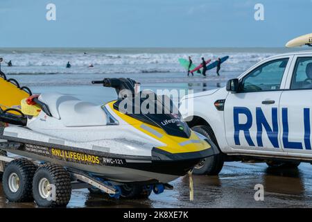 Un bagnino RNLI è in moto d'acqua e veicolo a Croyde Bay, North Devon, Regno Unito, con i surfisti che entrano in acqua sullo sfondo. Foto Stock