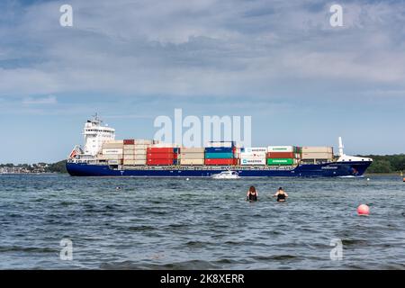 Kiel, Deutschland, Juli 2019 Ein Containerschiff fährt in die Kieler Förde am Falkensteiner Strand vorbei in die Kieler Innenförde, zwei badende fra Foto Stock