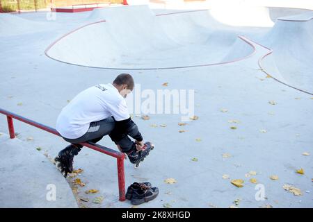 Giovane uomo in t-shirt bianca e jeans seduti sulla rampa e indossando le rollerblade. Allenamento nel fine settimana. Concetto di sport, hobby, stile di vita attivo Foto Stock