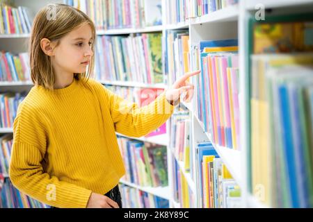 Libro di scelta della scolaretta nella biblioteca della scuola. Ragazza intelligente selezione letteratura per la lettura. Libri sugli scaffali in libreria. Imparare dai libri. Scuola ed Foto Stock