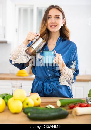 Giovane donna sorridente in vestito blu che versa caffè in tazza in cucina a casa Foto Stock