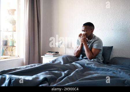 Spero che questa influenza vada via presto. Un bel giovane seduto nel suo letto e soffiando il naso mentre soffre di un freddo. Foto Stock