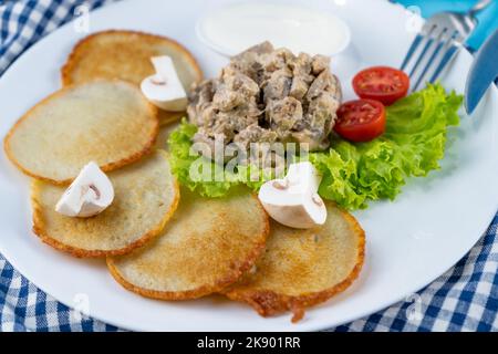 Deruny - frittelle di patate con funghi, champignons, panna acida su un piatto bianco. Pomodori ciliegini, insalata. Ristorante che serve su uno sfondo blu, tovagliolo a scacchi, forchetta, coltello. Vista dall'alto, spazio di copia Foto Stock