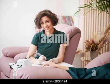 Ritratto del maestro di manicure che tiene la mano del cliente e che utilizza un trapano elettrico per unghie. Donna felice manicure guardando la macchina fotografica e sorridente mentre fa manicure per la donna in salone di bellezza. Foto Stock