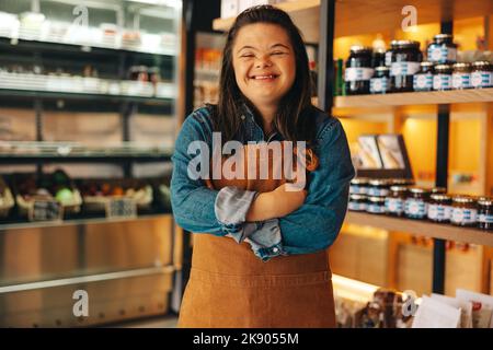 Operaio di negozio con sindrome di Down sorridente alla macchina fotografica mentre si trova in un negozio di alimentari. Donna abilitata con una disabilità intellettuale che lavora in un l Foto Stock