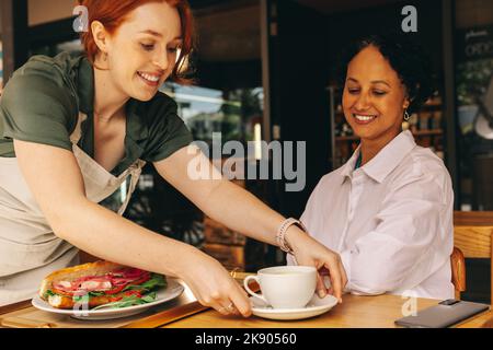 Cameriera giovane e felice che serve un panino e una tazza di caffè in un moderno caffè. Giovane donna allegra che lavora in un ristorante fast food. Foto Stock