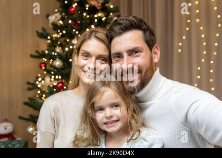 Ritratto di famiglia felice a Natale, famiglia uomo donna e figlia guardando la fotocamera e sorridendo, festeggiando il nuovo anno insieme a casa, utilizzando il telefono per la videochiamata e la comunicazione remota on-line. Foto Stock