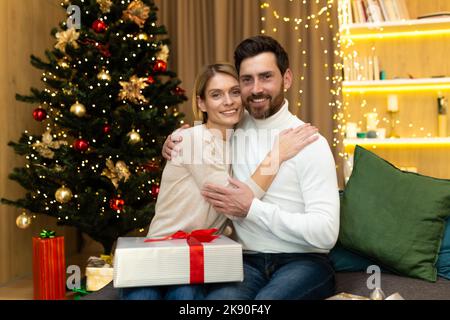 Ritratto di famiglia matura uomo bearded e donna bionda guardando la macchina fotografica che abbraccia vicino albero di Natale, coppia che celebra il natale e l'anno nuovo con i regali a casa seduto sul divano in soggiorno. Foto Stock