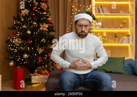 L'uomo è ammalato a casa, ha un grave mal di stomaco, siede sul divano vicino all'albero di Natale, è solo a casa per celebrare il nuovo anno in un cappello di Capodanno, tenendo lo stomaco con le mani. Foto Stock