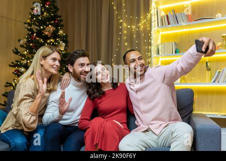 Festa di Capodanno a casa diversi gruppi di amici, uomini e donne, seduti sul divano, prendendo un selfie insieme, utilizzando uno smartphone, gli ospiti che guardano il telefono, parlando in una videochiamata per Natale Foto Stock