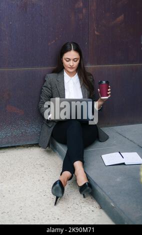 Vista frontale ritratto di una bella donna di successo seduta all'aperto ufficio, utilizzando computer portatile e bere caffè alla luce del mattino. Stile di vita e. Foto Stock