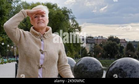 Attraente donna anziana mette mano a fronte, coprendo gli occhi dalla luce solare brillante, peering in distanza, cercando di vedere qualcuno. Elegante pensione Foto Stock