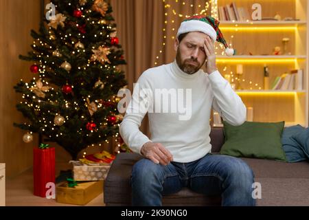 Uomo triste solo a Natale per due, seduto sul divano a casa in depressione vicino all'albero di Natale dopo aver festeggiato il nuovo anno. Foto Stock