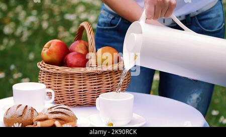 Sullo sfondo di un prato di camomilla, le mani delle donne versano il tè da una caraffa bianca in una tazza bianca, una bottiglia thermos. Sul tavolo c'è anche un cesto con mele rosse. Foto di alta qualità Foto Stock