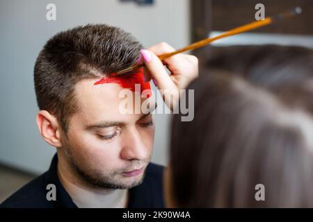 Ragazza artista dipinge su un viso mans. Pittura del viso sulla pelle. Ritratto di un giovane con vernice colorata su sfondo giallo. Trucco professionale F Foto Stock