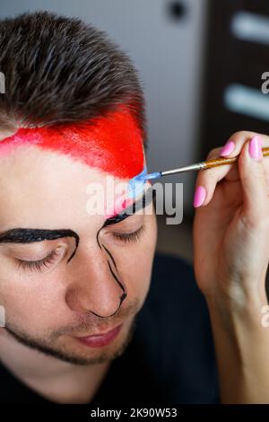 Ragazza artista dipinge su un viso mans. Pittura del viso sulla pelle. Ritratto di un giovane con vernice colorata su sfondo giallo. Trucco professionale F Foto Stock