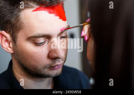 Ragazza artista dipinge su un viso mans. Pittura del viso sulla pelle. Ritratto di un giovane con vernice colorata su sfondo giallo. Trucco professionale F Foto Stock