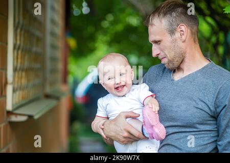 padre tiene in braccio la figlia di un anno. L'amore di Daddy per la ragazza. Il bambino si diverte all'aperto il giorno d'estate. Attività di famiglia. Il bambino sorride Foto Stock