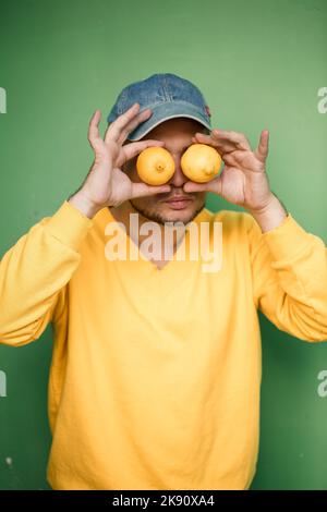 Giovane con la barba in un cappello sulla testa con i limoni sugli occhi su uno sfondo verde. Ritratto di un ragazzo in un maglione giallo Foto Stock