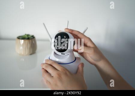 Primo piano sulla sezione centrale e sulle mani di una donna caucasica sconosciuta che tiene in mano la telecamera di sorveglianza di sicurezza domestica mentre si è in piedi in sala per la regolazione e la configurazione Foto Stock