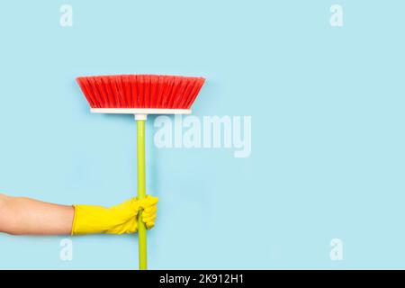 Mano donna con guanto di gomma gialla che tiene una scopa rossa su sfondo azzurro con spazio copia Foto Stock