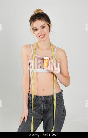 giovane donna con figura sottile in piedi con metro a nastro e vitamine isolato sul grigio Foto Stock