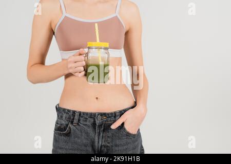 vista ritagliata di una giovane donna in forma con frullato mentre si è in piedi con la mano in tasca isolato sul grigio Foto Stock