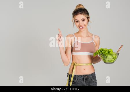 donna sorridente con metro a nastro in vita tenendo lattuga e mostrando idea gesto isolato sul grigio Foto Stock