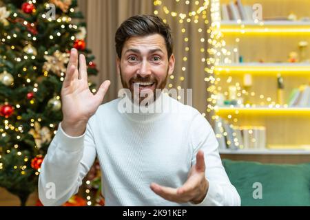 Primo piano uomo felice che parla con una videochiamata guardando webcam e parlando con gli amici, a casa per Natale seduto su un divano nel salotto vicino all'albero di Natale, nelle vacanze di Capodanno. Foto Stock