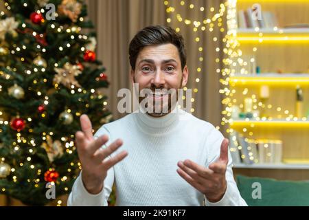 Primo piano uomo felice che parla con una videochiamata guardando webcam e parlando con gli amici, a casa per Natale seduto su un divano nel salotto vicino all'albero di Natale, nelle vacanze di Capodanno. Foto Stock