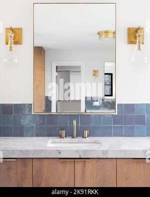 Una casa di lusso rinnovato bagno con piastrelle blu, un mobile di vanity in legno chiaro, luci dorate montate sulla parete, e un banco di pietra grigio. Foto Stock