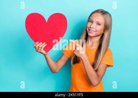 Foto di bella studentessa bambino indossare t-shirt arancione sorriso toothy tenere grande carta rossa cuore dito puntando sentimenti prima data isolato sul colore ciano Foto Stock