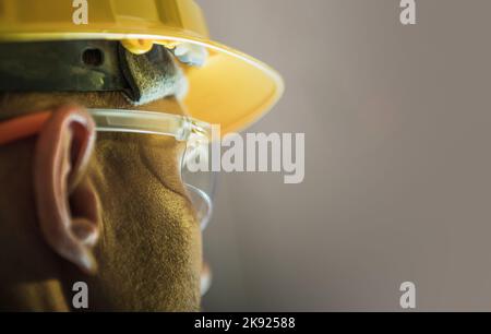 Primo piano di un lavoratore caucasico maschio che indossa un cappello giallo duro e occhiali protettivi. Sfondo solido con spazio di copia. Tema dell'equipaggiamento di sicurezza per la costruzione. Foto Stock