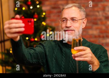 Un uomo anziano in una maglia verde che parla con la famiglia dal collegamento video del telefono vicino all'albero di Natale Foto Stock