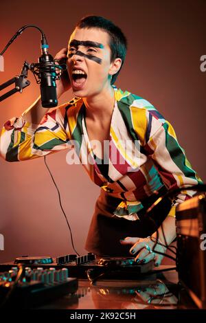 Musicista che urla al microfono, mixando musica techno ai giradischi dj su sfondo fumoso. Artista femminile che suona suoni a strumenti audio e mixer stereo. Foto Stock