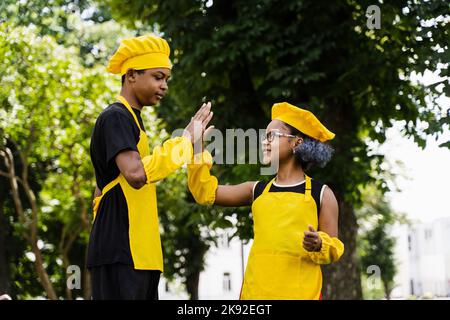 l'africa nera cucina i bambini in cuochi gialli cappello e grembiule si battono le mani e danno cinque. Fratello africano nero e sorella che cucinano e che hanno f Foto Stock