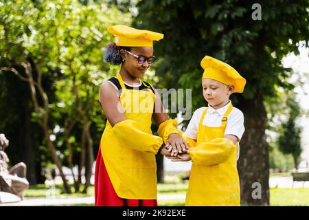 Comunicazione tra amici multirazziali di neri africani e caucasici cuoco bambini in giallo chef cappello e grembiule tenendo le mani e divertirsi Toge Foto Stock