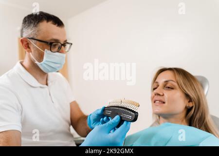 Il dentista dimostra le sfumature di colore dei denti guida per sbiancare i denti per la paziente donna in clinica dentale. Odontoiatria. Donna che guarda impiallacciature o impianti t Foto Stock