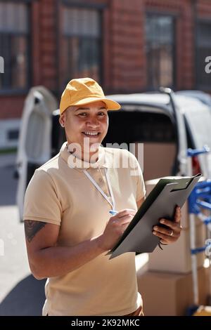 Ritratto verticale del sorridente addetto alla consegna che tiene la scatola degli appunti mentre è in piedi da un furgone alla luce del sole Foto Stock