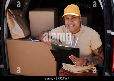 Ritratto di donna sorridente come lavoratore di consegna che guarda la fotocamera mentre scrive sulla clipboard in furgone tronco, copia spazio Foto Stock