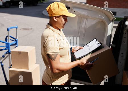 Vista laterale ritratto di giovane donna multietnica che scarica furgone con pacchi mentre lavora nel servizio di consegna Foto Stock