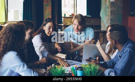 Giovani colleghi di sesso maschile e femminile stanno esaminando i documenti che si trovano sul tavolo e parlano condividendo idee. Concetto di lavoro di squadra, ufficio moderno, informazioni e dipendenti aziendali. Foto Stock