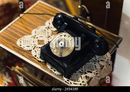 Elegante telefono vintage autentico nero lucido con quadrante rotondo sdraiato su un piccolo tavolo di legno con fancywork decorativo fatto a mano. Accessori per la casa Hippster vintage. Illustrazione di alta qualità Foto Stock