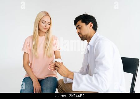 Studio shot di grave chirurgia del medico maschile in uniforme esame mano rotta avvolto in cerotto di cerotto di paziente femminile blonde ferito a. Foto Stock