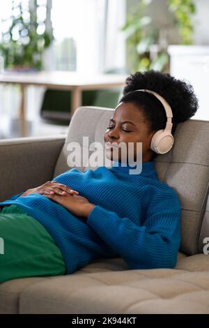 Donna afro-americana riposante in cuffie con un lieve sorriso giace sul divano addormentato alla musica Foto Stock