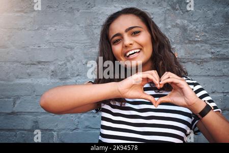 Il mio cuore è pieno di amore per te. Ritratto di una giovane donna a forma di cuore con le mani contro una parete grigia. Foto Stock