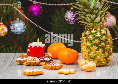 Ananas, mandarini e un abete con ornamenti natalizi sullo sfondo. Foto Stock