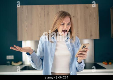 Donna arrabbiata confusa che ha problemi con il telefono in cucina a casa, giovane donna infelice che guarda lo schermo, insoddisfatto per dimesso o rotto Foto Stock