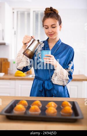 Donna in accappatoio blu che versa caffè vicino al tavolo con muffin al forno Foto Stock