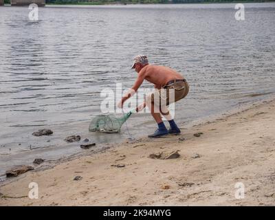 Un vecchio uomo in pantaloncini corti e un cappello e un cavalcetto di pesca in mano che tira il pesce fuori dal fiume Dnipro su una spiaggia urbana vicino al ponte Nord nella città di Kyiv. Foto Stock